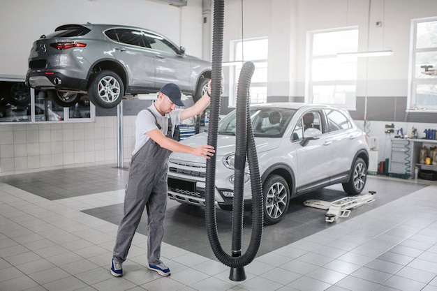 Werknemer in uniforme stand en houd lange buis met beide handen vast. Hij kijkt naar beneden. Jonge man is serieus en geconcentreerd. Er zijn twee auto's achter hen. Een ervan staat op het platform.