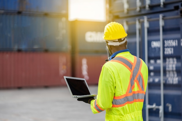 Foto werknemer man in beschermende veiligheid jumpsuit uniform met gele veiligheidshelm en laptop check container gebruiken bij verzending vrachtmagazijn. transport import, export logistieke industriële service