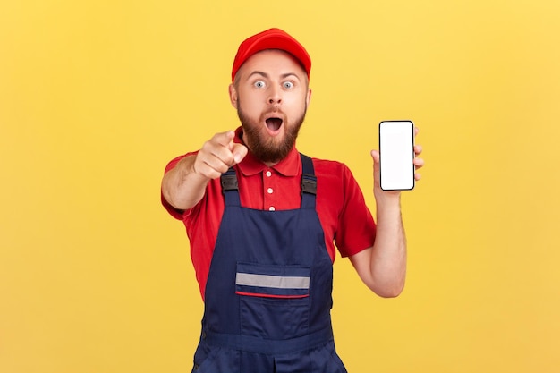 Werknemer man in blauwe overall t-shirt wijzende vinger naar jou en toont smartphone met leeg display