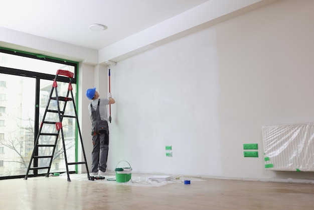 Werknemer met helm schildert muur met rolborstel in ruime premisse bouwer renoveert kamer