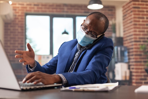 Werknemer praten over telefoongesprek met behulp van mobiele telefoon en werken op laptop tijdens pandemie. zakenman met gesprek op smartphone om project en strategie te plannen terwijl hij gezichtsmasker draagt.