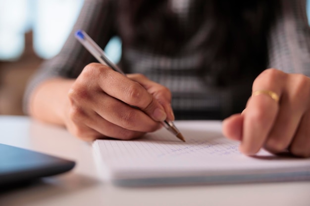 Werknemer schrijft informatie op notebook, maakt aantekeningen over bedrijfsrapport over leerboek thuis. Werken aan het opstarten van papierwerk en het bijwonen van een online klaswebinar op de computer. Detailopname.