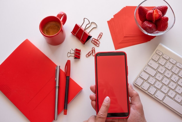 Werknemer van huis heeft een pauze met koffie en aardbeien, met behulp van telefoon met rood scherm. Witte desktop en rode accessoires