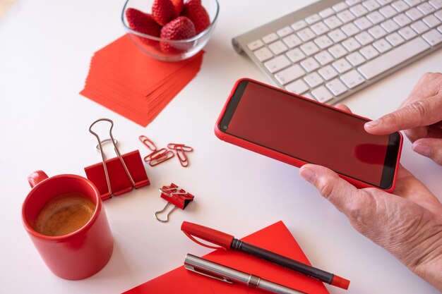 Werknemer van huis heeft een pauze met koffie en aardbeien, met behulp van telefoon met rood scherm. Witte desktop en rode accessoires