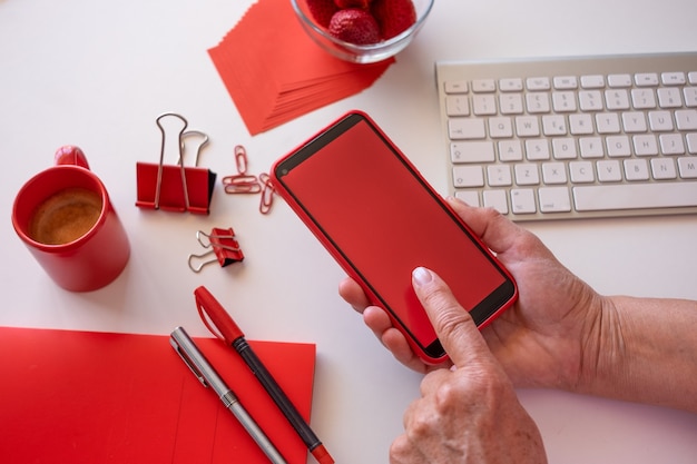 Werknemer van huis heeft een pauze met koffie en aardbeien, met behulp van telefoon met rood scherm. Witte desktop en rode accessoires