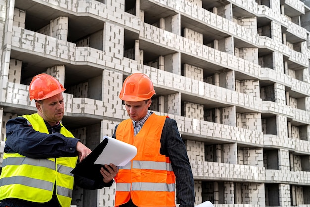 Werknemers in uniform en helmen vergelijken het bouwplan bij het nieuwe gebouw op een tablet. Het concept van een nieuw gebouw