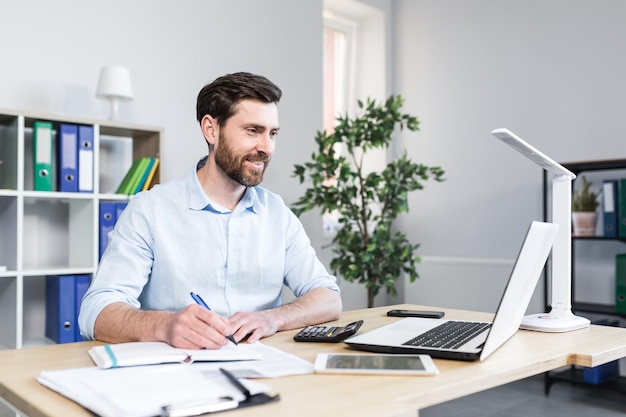 Werkomgeving op kantoor Portret van een jonge mannelijke zakenman-econoommanager die op kantoor aan het bureau werkt op een laptop die met papieren documenten werkt