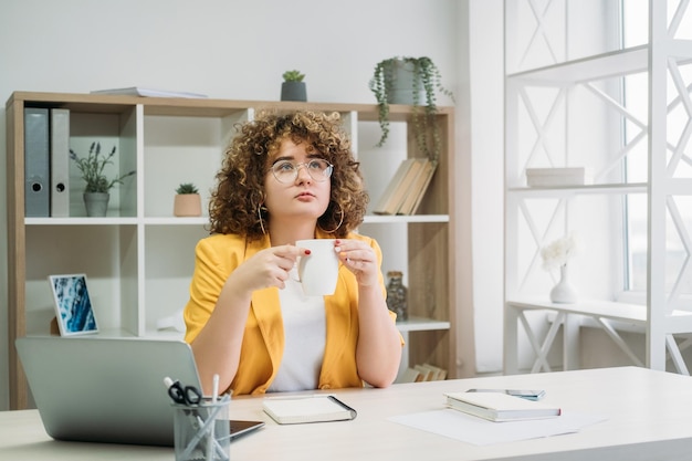 Werkpauze Koffietijd Externe baan Doordachte peinzende overgewicht krullend haar vrouw werknemer met laptop met kopje op licht kantoor aan huis