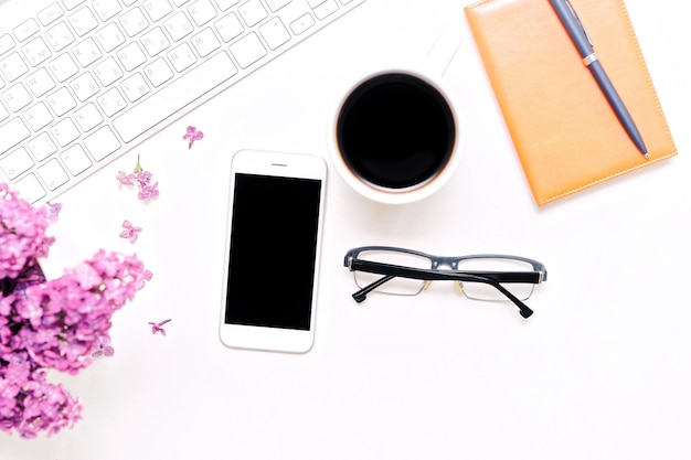 Werkplek met computertoetsenbord, mobiele telefoon, kopje koffie, notebook, pen, bril en lila bloemen op de witte tafelachtergrond. Platliggend, bovenaanzicht