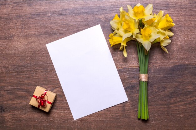 Werkplek met een cadeau met een rood lint, een stuk papier en een boeket narcissen op een houten achtergrond. Plat ontwerp, bovenaanzicht.