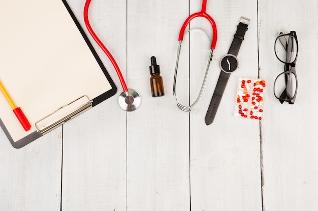 Werkplek van een arts Rode stethoscoop klembord bril horloge en pillen op witte houten tafel