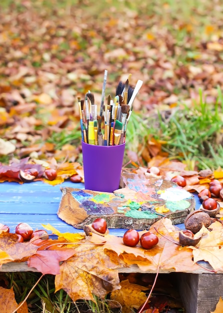 Werkplek voor schilder in herfstpark met houtpalet en penselen in plastic container. herfstbladeren en kastanjes op blauwe houten achtergrond.