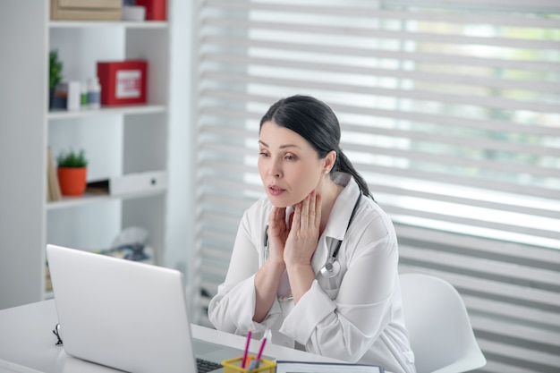 Werkproces. Succesvolle mooie vrouw in een wit gewaad zit een laptop zorgvuldig kijken naar het scherm in haar kantoor.