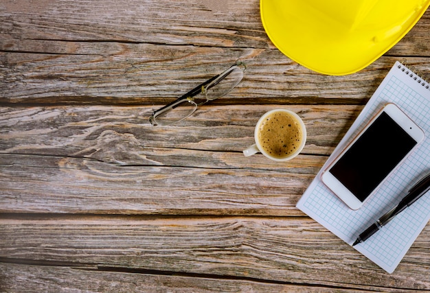 Werkruimte bouwers architect bureau, lege notitieblok openen met pen op gele helm met kopje koffie, smartphone brillen op houten bureau