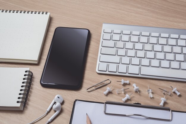 Werkruimte met dagboek of notebook en klembord, muiscomputer, toetsenbord, smartphone, oortelefoon, potlood, pen op hout