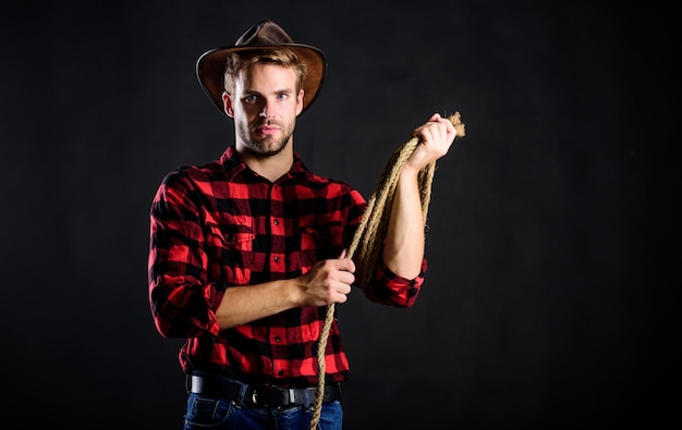 Western life Man unshaven cowboy black background Man wearing hat hold rope Lasso tool of American cowboy Lasso used rodeos competitive events Lasso can be tied or wrapped Brutal and confident