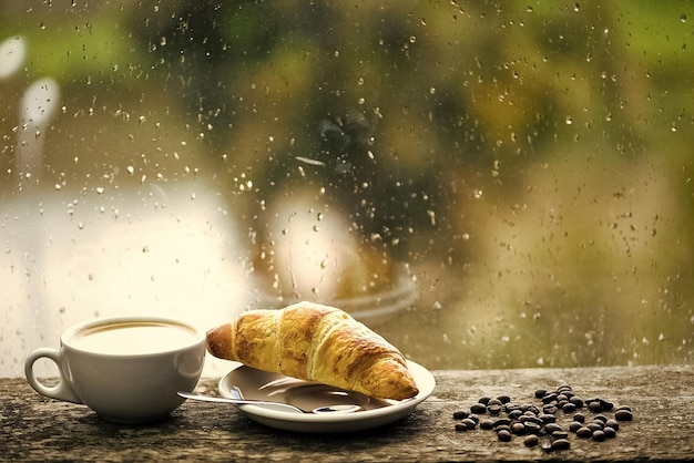 Wet glass window and cup of hot caffeine beverage coffee drink\
with croissant dessert enjoying coffee on rainy day coffee time on\
rainy day fresh brewed coffee in white cup or mug on\
windowsill