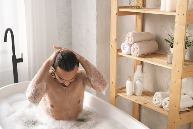 Bagnato giovane uomo muscoloso lavarsi i capelli mentre si fa il bagno con schiuma da scaffali con oggetti di auto-amore