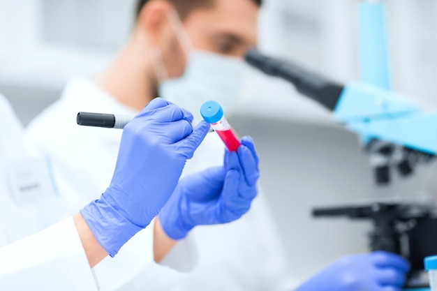 wetenschap, scheikunde, technologie, biologie en mensen concept - close-up van wetenschappers handen schrijven op reageerbuis met marker en onderzoek doen in klinisch laboratorium