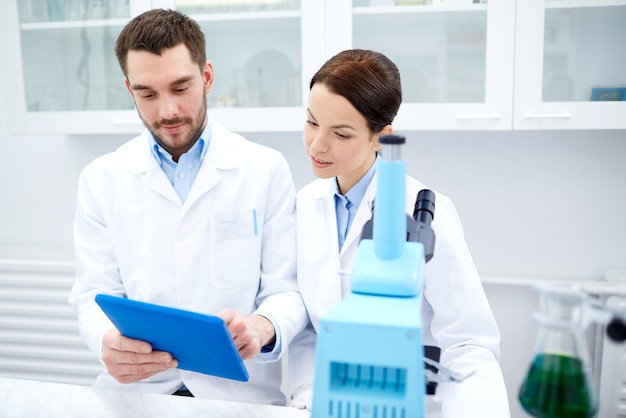 wetenschap, scheikunde, technologie, biologie en mensenconcept - jonge wetenschappers met tablet-pc en microscoop die test of onderzoek maken in klinisch laboratorium