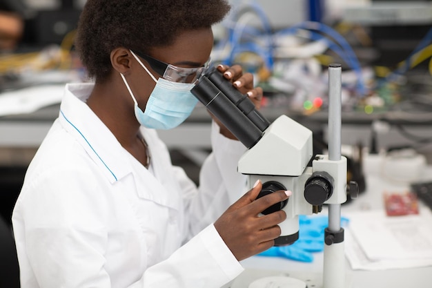 Wetenschapper afro-amerikaanse vrouw in gezichtsmasker en handschoenen werken in laboratorium met elektronische technische instrumenten en microscoop Onderzoek en ontwikkeling van elektronische apparaten door kleur zwarte vrouw