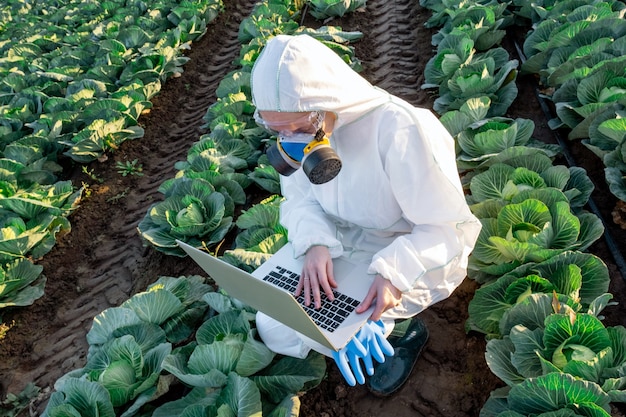 Wetenschapper die een witte beschermende uitrusting, een chemisch masker en een bril draagt, gebruikt laptop op het gebied van de boerderij.