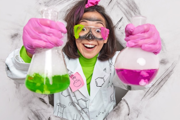 Foto wetenschapper houdt twee kolven vast met groene en roze vloeistof toont resultaten van chemisch onderzoek draagt vuile veiligheidsbril en jas breekt door papier