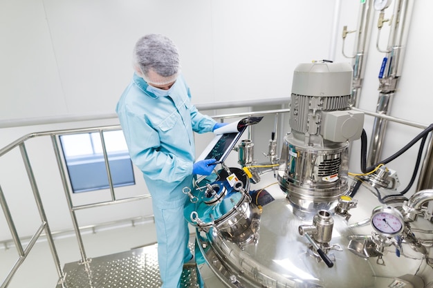 Wetenschapper in blauw laboratoriumpak en gasmasker staan in de cleanroom en controleren grafieken in tablet in de buurt van grote stalen tank