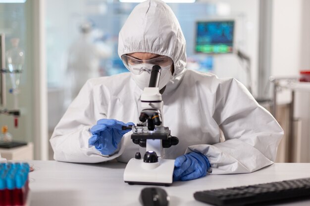 Wetenschapper in pbm-pak die aanpassingen maakt en door laboratoriummicroscoop kijkt