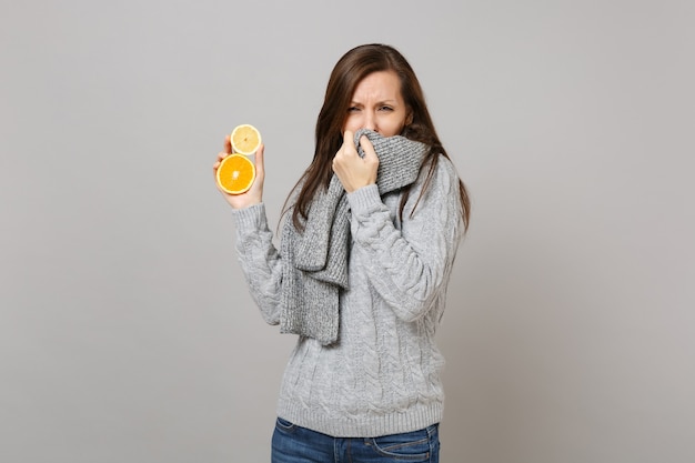 Whacked jonge vrouw in grijze trui die mond bedekt met sjaal, niezen of hoesten, houd citroen, sinaasappel geïsoleerd op een grijze achtergrond. gezonde levensstijl, behandeling van zieke ziektes, concept van het koude seizoen.