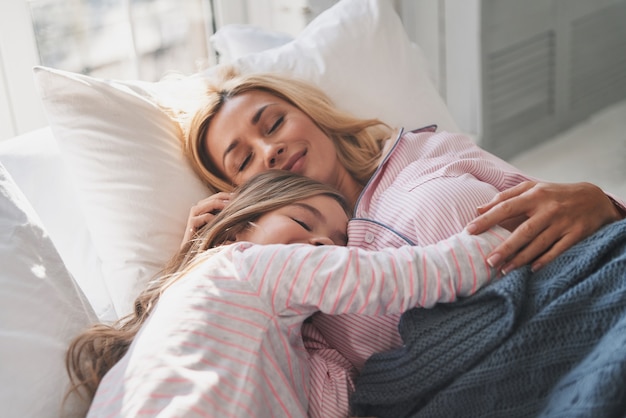 What are they dreaming about? Mother and daughter keeping eyes closed and smiling while lying on the bed at home