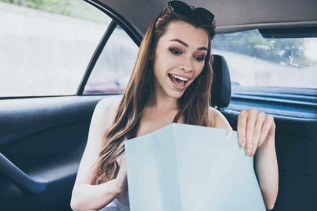 What a beautiful dress! Beautiful young smiling woman looking inside of the shopping bag and looking surprised while sitting inside of the car