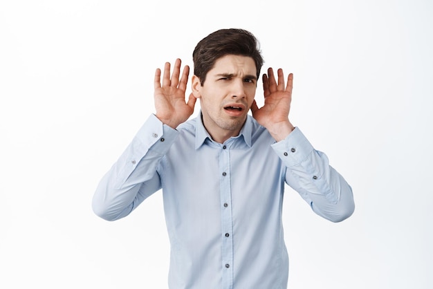 What I cant hear you Confused male office manager holding hands near ears and looking puzzled listening closer trying to eavesdrop standing against white background