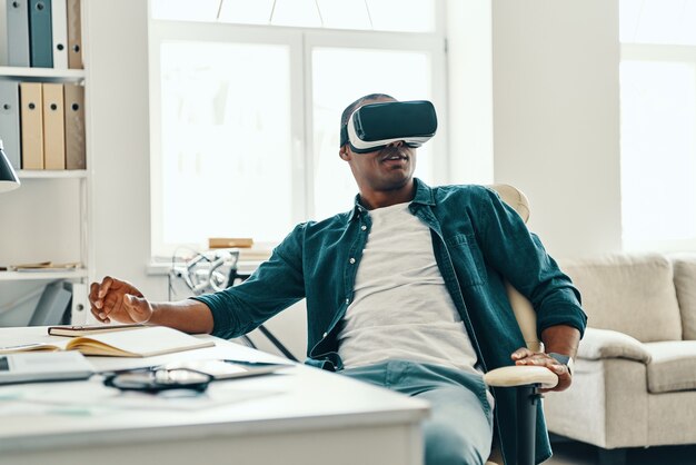 What is real? Handsome young African man wearing virtual reality simulator headset while sitting indoors