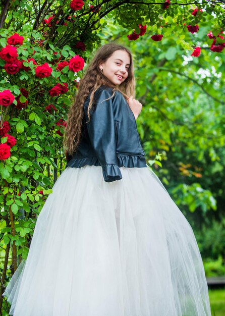 What a lovely dress. beauty and fashion. pretty kid smell rose flower. spring and summer nature. little girl in garden. child enjoy blossom in park. small cute lady in leather jacket. happy childhood.