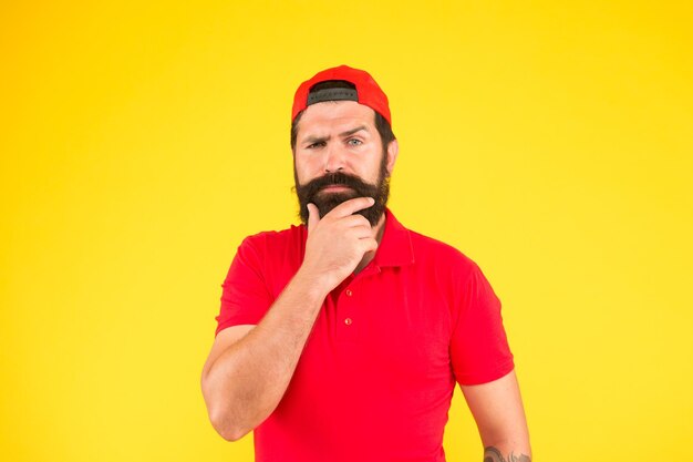 What shampoo to choose being trendy and brutal Beard grooming thoughtful mature hipster yellow background bearded man red shirt and cap male summer fashion Barber salon and facial hair care