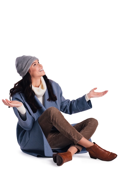 What a surprise! Beautiful young cheerful woman in beany keeping arms outstretched and looking up with smile while sitting against white background