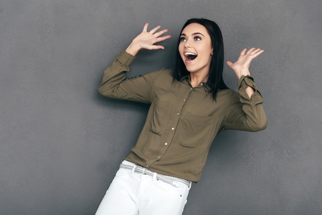 What a surprise! Surprised young smiling woman in smart casual wear standing against grey background and gesturing