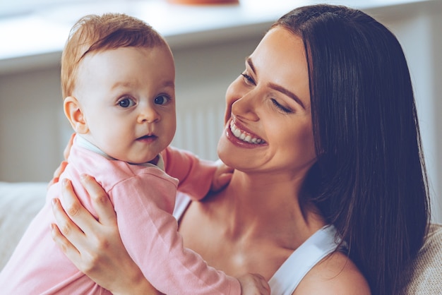 What a sweet cutie! Cheerful beautiful young woman holding baby girl in her hands and looking at her with love while sitting on the couch at home