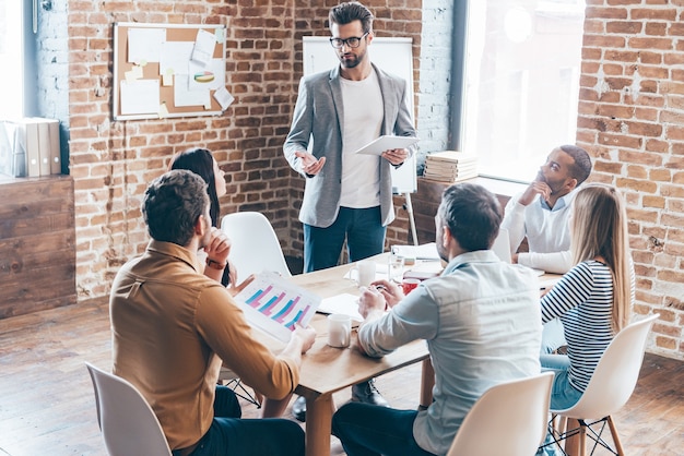 What do you think about it? Young handsome man wearing glasses holding digital tablet and discussing something while his coworkers listening to him sitting at the office table