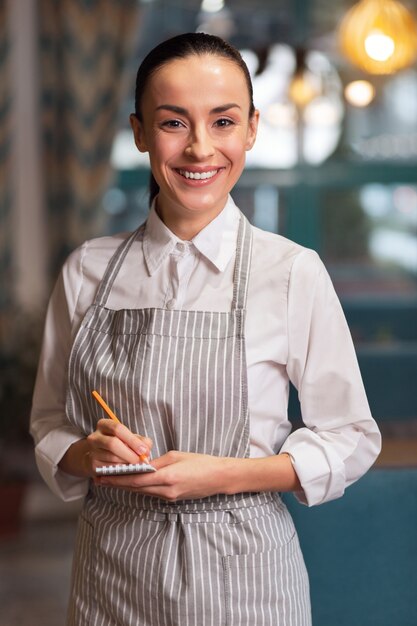 What you want. Brunette nice cheerful waitress holding notebook while looking at camera and smiling