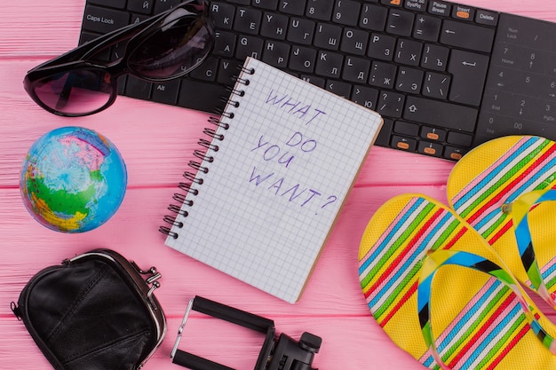 What do you want on notebook with woman\'s traveler accessories\
glasses wallet and flip-flops on pink table top background.globe\
and black keyboard.