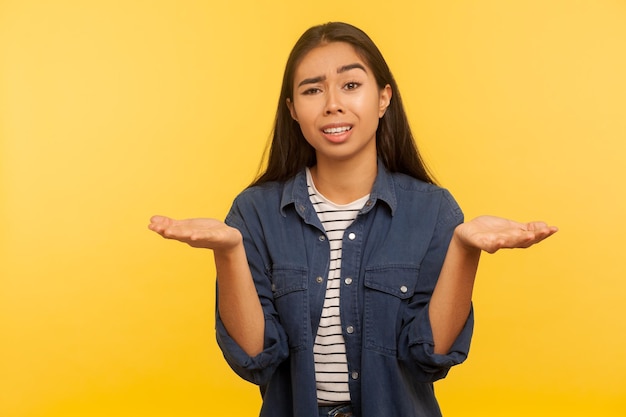 What do you want. portrait of confused frustrated girl in denim\
shirt shrugging shoulders with indignant expression, asking why\
misunderstanding. indoor studio shot isolated on yellow\
background