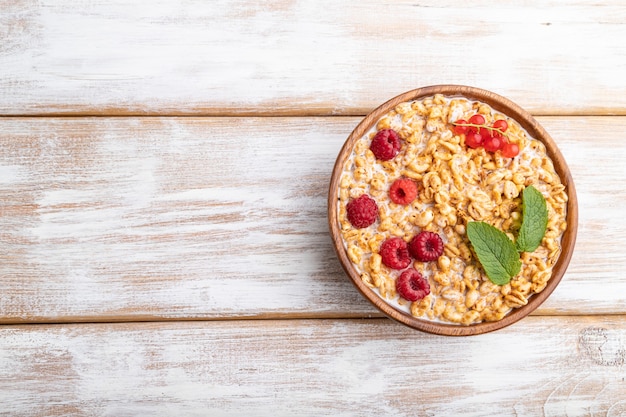 Wheat flakes porridge with milk, raspberry and currant in wooden bowl on white wooden background. top view, flat lay, copy space