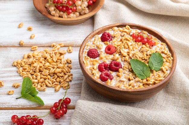 Wheat flakes porridge with milk, raspberry and currant in wooden bowl on white wooden surface and linen textile