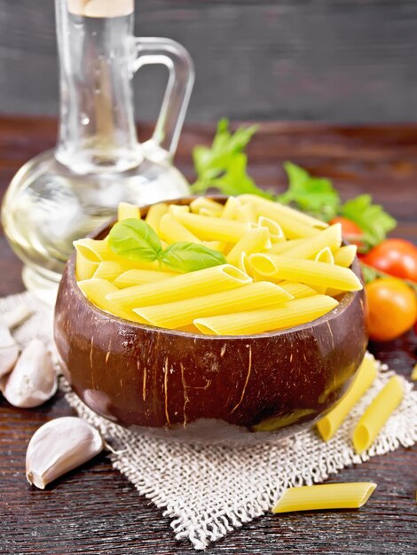 Wheat flour penne pasta in a bowl of coconut shells on sacking tomatoes garlic vegetable oil in a decanter and parsley on wooden board background