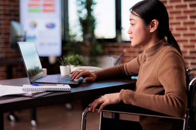 Wheelchair user planning business project on laptop browser, dealing with chronic health condition. Paralyzed woman with physical impairment working in disability friendly office.