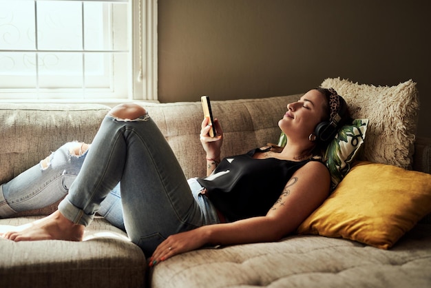 Photo when words are few music says a lot shot of a young woman using a smartphone and headphones on the sofa at home