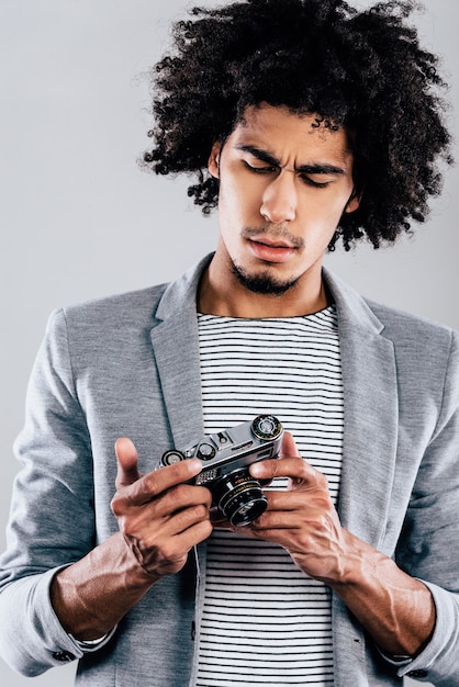 Where is digital screen here? Handsome young African man holding retro styled camera and looking at it while standing 