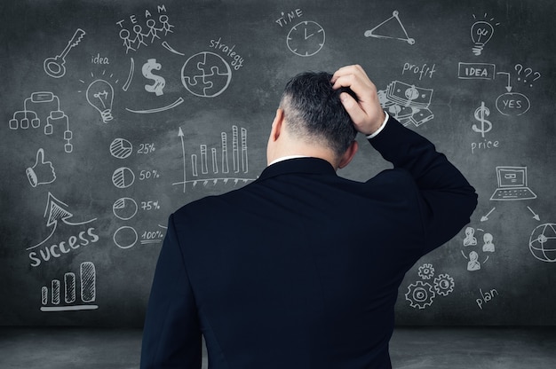 Which way shall I choose? Rear view of pensive mature man scratching his head and looking at digitally composed business signs and charts on chalkboard